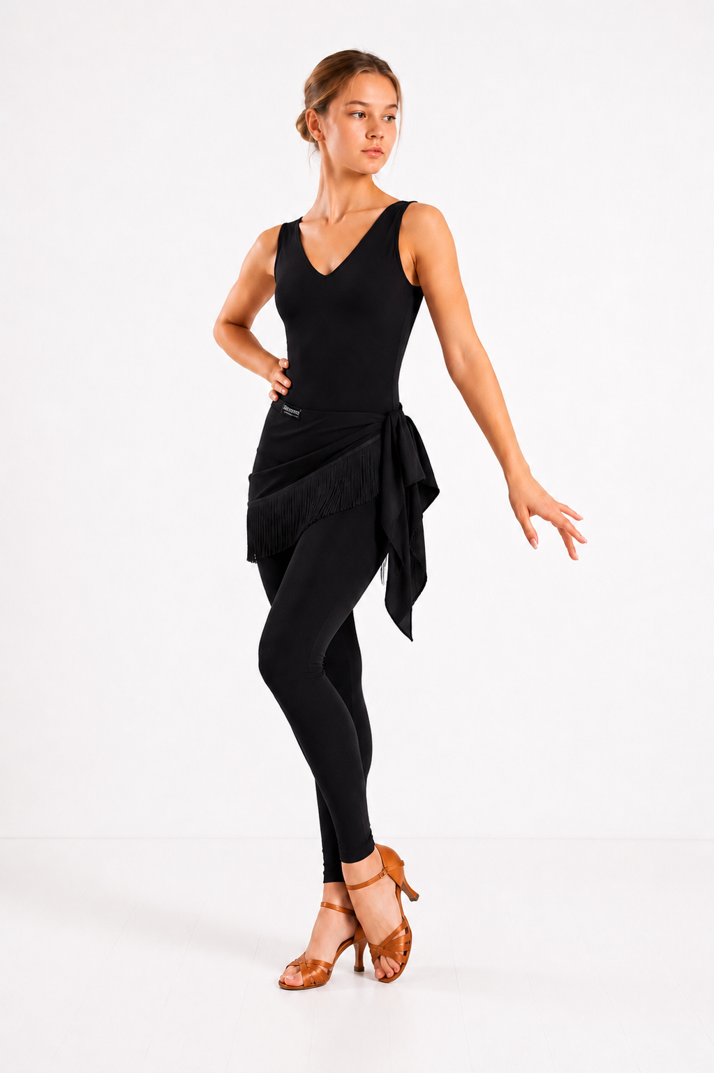 Woman wearing a black Latin dance jumpsuit with a black fringe hip scarf wrap skirt and tan dance shoes on a white studio background.